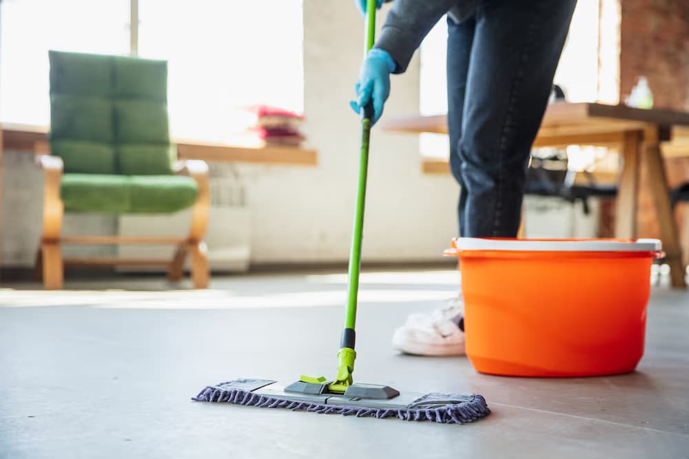 Person mopping a floor with a green mop and orange bucket in a bright interior space.